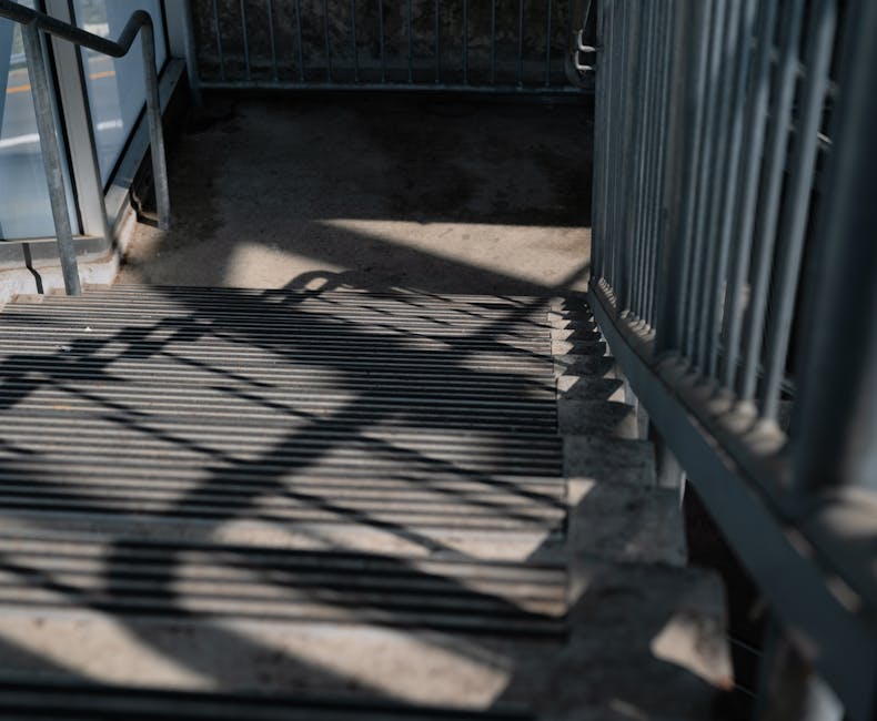 Close-up view of outdoor metal stairs with horizontal slats, showing shadows cast by the railing on the concrete ground and stair surface. The stairs are part of an entrance to a residential building,