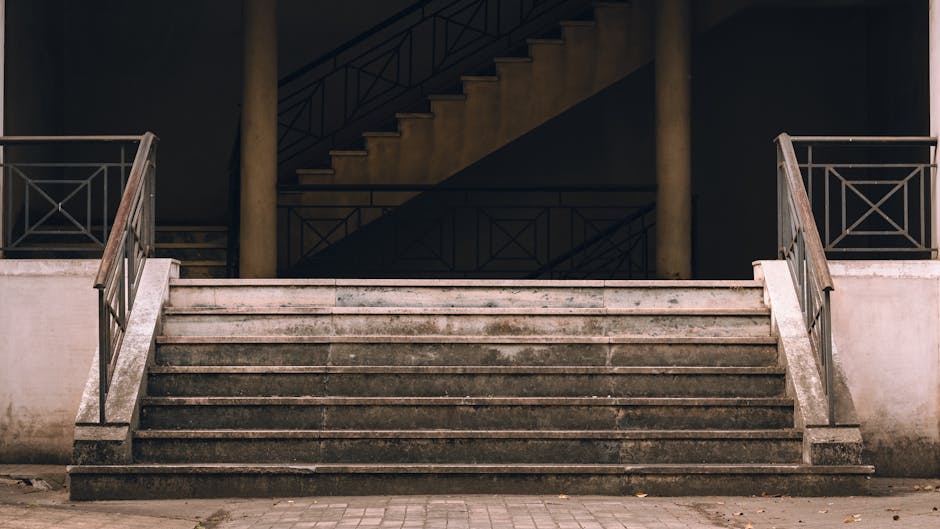 Close-up view of outdoor metal stairs with horizontal slats, showing shadows cast by the railing on the concrete ground and stair surface. The stairs are part of an entrance to a residential building,