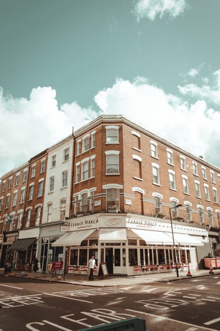 An exterior view of a corner building with a ground-floor café named 'Franko Manca,' featuring a white awning and large glass windows. The building is made of red brick with white-trimmed windows, positioned on a street corner in Finchley. The street has road markings and a pedestrian crossing, with a few people walking nearby. Bright daylight and partly cloudy skies cast natural light over the scene, creating a clean and inviting atmosphere indicative of professional cleaning and maintenance. Cleaner Finchley specializes in domestic and commercial surface cleaning, including exterior surface sanitisation and deep cleaning treatments, ensuring buildings like this are kept presentable and hygienic.