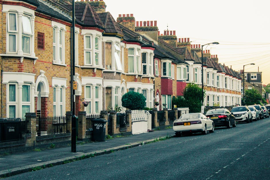 A row of traditional British terraced houses with brick facades and white bay windows lining a residential street in Finchley. The pavement is clean and well-maintained, with small front gardens enclosed by low brick walls and black metal railings. Several parked cars are aligned along the street, varying in size and color, including white, black, and silver vehicles. The street lighting and overhead power lines are visible under an early evening sky, casting a soft light over the scene. The overall appearance of the neighborhood suggests a tidy and well-kept residential area, ideal for domestic or deep cleaning services. This image is associated with Cleaner Finchley's guidance on flat cleaning for renters in Finchley, emphasizing property upkeep and hygiene.