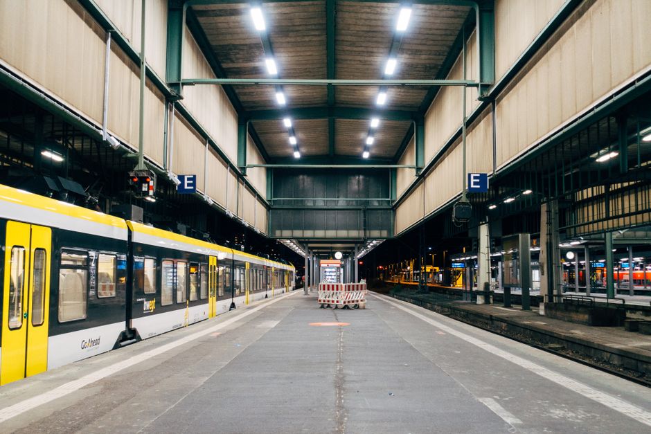 A well-lit train station platform with a bright yellow and white commuter train stopped on the left side. The platform surface is clean, made of concrete with a textured safety strip along the edge. Overhead lighting illuminates the area, casting light on the adjacent walls and ceiling, which are constructed from wood and metal with visible support beams. The station features multiple tracks on the right with additional trains and station facilities in the background. The overall appearance emphasizes a tidy, maintained environment, reflecting thorough surface cleaning and hygiene. Cleaner Finchley offers professional cleaning services that include deep cleaning and sanitisation for transportation hubs like this, ensuring safe and hygienic spaces.