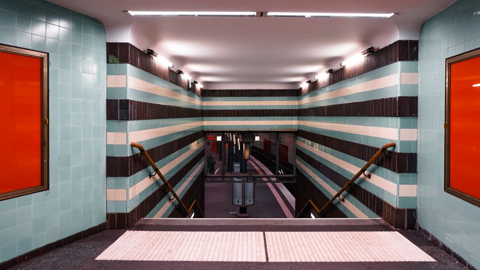 A tiled underground station entrance with turquoise, white, and black horizontal striped ceramic wall tiles. The image shows a descending staircase with wooden handrails on both sides, leading to an area with a tiled floor and additional architectural features in the distance. The lighting is bright and even, highlighting the clean, polished surfaces of the tiles and the tidy appearance of the space, indicative of a professional deep cleaning process by Cleaner Finchley. The station's window frames are orange, contrasting with the cool tones of the tiles, and the overall scene reflects hygiene and maintenance suitable for a public transportation environment.