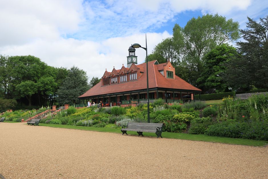 Exterior view of Stephens House and Gardens showing a red brick building with Gothic-style arched windows framed in white stone, surrounded by lush greenery and shrubs. The foreground features a grave
